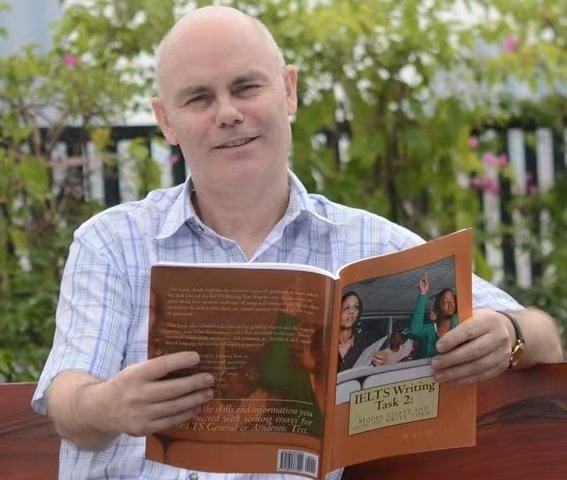 Mike Wattie, former IELTS examiner, holding a co-authored IELTS Writing Task 2 textbook